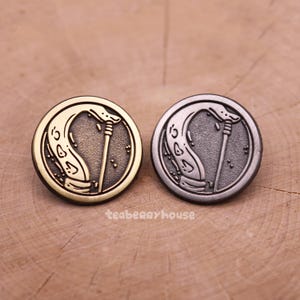 May include: Two circular metal buttons featuring a snake and staff design. One button is gold-coloured, the other is silver-coloured. The buttons have a vintage aesthetic. The text "teaberryhouse" is visible on the wooden surface.