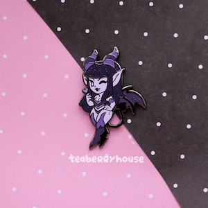 Puede incluir: Un pin de esmalte morado y negro con una chica demonio de dibujos animados con cuernos, alas y un corazón. El pin está sobre un fondo de lunares rosas y negros. El texto "teaberryhouse" es visible.