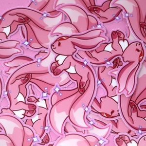 Może przedstawiać: Stos różowych i białych naklejek przedstawiających kreskówkowego lisa z puszystym ogonem. Naklejki mają błyszczący, migoczący efekt.