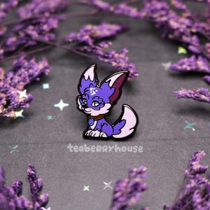 Puede incluir: Pin de esmalte morado y blanco de un zorro de dibujos animados con una gema azul en su collar. El zorro tiene un patrón de remolino morado y blanco en su pelaje y un ojo azul con una estrella blanca.