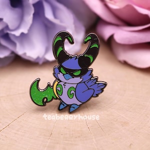 May include: A colorful enamel pin featuring a cartoon owl-like creature with black horns and a green, glittery wing. The creature has a blue body, a black mask, and green eyes. The pin has the text "teaberryhouse".