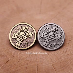 May include: Two round metal pins, one gold-toned and one silver-toned, featuring a stylized skeletal hand design within a circle. The pins have a detailed, engraved appearance with star accents. The text "teaberryhouse" is printed on the wooden surface.