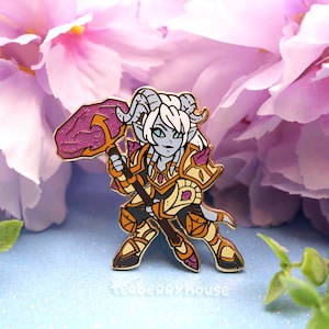 Könnte beinhalten: Eine goldfarbene Emaille-Anstecknadel mit einer Cartoon-Figur mit violetten Hörnern, blauer Haut und weißen Haaren. Die Figur trägt eine goldene Rüstung und hält eine violette und goldene Waffe. Die Anstecknadel ist vor einem Hintergrund aus rosa Blumen platziert.