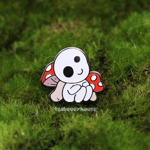 May include: A whimsical enamel pin featuring a white creature with a mushroom cap. The mushroom has red caps with white spots. The pin is set against a backdrop of green moss.