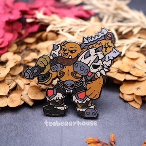 May include: A brown and black enamel pin of a cartoon orc warrior holding a gun and wearing a skull belt. The orc has a beard and is wearing armor. The pin is on a gray background.
