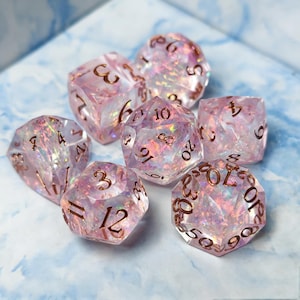 Può includere: Un set di sette dadi poliedrici realizzati in resina rosa traslucida con un effetto lamina interno iridescente e scintillante. Ogni dado presenta numeri incisi riempiti con inchiostro metallico color rame, inclusi un d20, d12, d10, d8, d6, d4 e un dado percentuale.