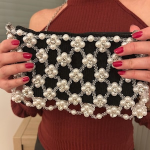 Peut inclure: Pochette noire avec un motif floral en perles blanches et perles transparentes. La pochette a une bandoulière en chaîne argentée et une fermeture éclair. Le motif floral est orné de petits strass étincelants.
