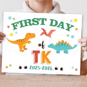 Puede incluir: Un letrero blanco con el texto "FIRST DAY of TK 2025-2026" en verde, rojo y naranja. El letrero presenta dinosaurios de dibujos animados en naranja y azul, con estrellas y huellas. El letrero está sostenido por una persona.