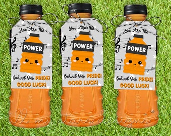 Op de afbeelding: Drie oranje sportdrankflessen met zwarte doppen en aangehechte labels. De labels lezen "You Are The POWER" met een cartoon fles grafiek en muzieknoten. Tekst op de labels zegt "Behind Our PRIDE! GOOD LUCK!"