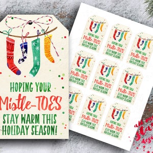 May include: A set of 10 Christmas gift tags featuring a watercolour illustration of three colourful socks hanging on a clothesline. The tags have the text "Hoping your Mistle-Toes Stay Warm This Holiday Season!" printed on them.