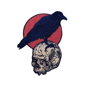 May include: Embroidered patch featuring a black crow perched atop a skull. The crow is set against a red circle, with the skull in a beige color. The patch has a gothic or dark aesthetic.