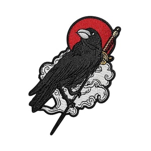 May include: Embroidered patch featuring a black crow perched on a cloud with a sword. The crow is set against a red sun, with gold and red accents on the sword. The patch has a white background.