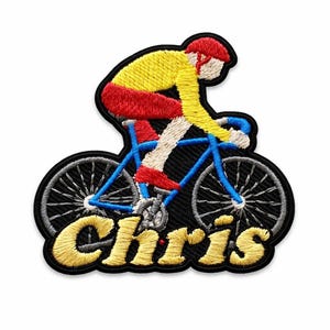 May include: Embroidered patch featuring a cyclist in yellow and red riding a blue bicycle. The patch has the name "Chris" in yellow script. The patch is outlined in black.