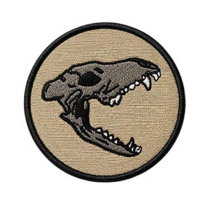 May include: Embroidered patch featuring a detailed illustration of a canine skull with visible teeth. The skull is gray, set against a tan background, and framed by a black border. The patch is round and suitable for jackets, bags, or other fabric items.