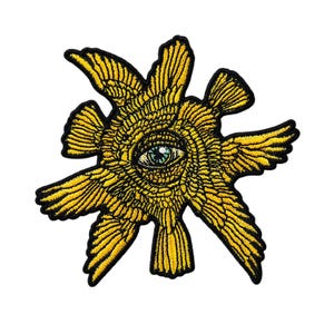 May include: Embroidered patch featuring a stylized eye within a sunburst design. The patch has a yellow and black color scheme, with detailed wing-like elements radiating outwards. The eye is detailed with blue and green accents.