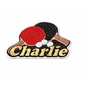 Könnte beinhalten: Gestickter Aufnäher mit zwei Tischtennisschlägern, einem roten und einem schwarzen, mit drei weißen Tischtennisbällen. Der Aufnäher hat den Namen "Charlie" in goldfarbenen Buchstaben mit schwarzer Umrandung. Der Aufnäher befindet sich auf weißem Hintergrund.