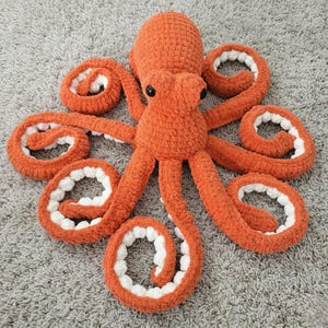 Può includere: Un peluche a forma di polpo all'uncinetto arancione fatto a mano con dettagli bianchi sui tentacoli. Il peluche ha occhi neri a bottone ed è appoggiato su un tappeto grigio. Questo giocattolo morbido è adatto ai bambini.