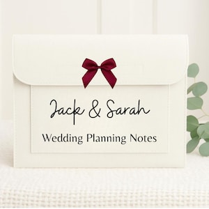 Peut inclure: Pochette de notes de planification de mariage de couleur crème avec un nœud bordeaux. Le dossier a une fermeture à rabat et une poche avec les noms "Jack & Sarah" et le texte "Wedding Planning Notes" en script noir.