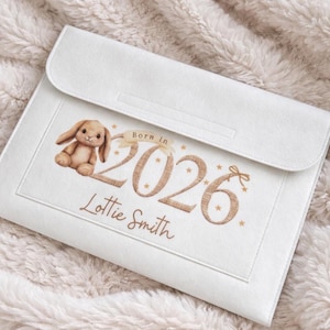 May include: White baby keepsake case with a flap closure. The case features a brown bunny illustration, the text "Born in 2026", and the name "Lottie Smith" in a script font. The case is on a soft, textured surface.