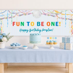 May include: A rectangular party banner with a light blue and white vertical stripe background, featuring a disco ball, colorful streamers, and the text "FUN TO BE ONE! Happy Birthday Preston!" displayed above a table with a cake and gifts.