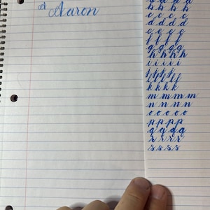 May include: A spiral-bound notebook with blue-lined paper. The name "Aaron" is written in cursive at the top. A separate sheet displays cursive letter practice in blue ink, including the letters y, b, c, d, e, f, g, h, i, j, k, m, n, p, q, and s.