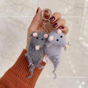 Puede incluir: Dos llaveros de ratón hechos a mano. Uno es gris, el otro es lavanda. Ambos tienen orejas y narices rosas, ojos negros y bigotes. Los llaveros están unidos a un anillo plateado.