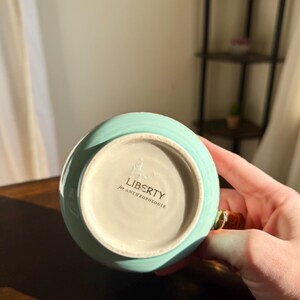 Puede incluir: Una taza de cer&aacute;mica turquesa con un asa dorada. La base de la taza tiene el texto "LIBERTY for ANTHROPOLOGIE". La taza est&aacute; sujeta por una persona.