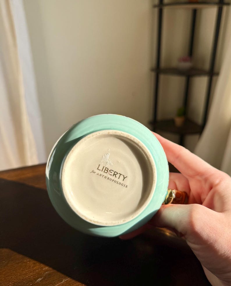 Puede incluir: Una taza de cer&aacute;mica turquesa con un asa dorada. La base de la taza tiene el texto "LIBERTY for ANTHROPOLOGIE". La taza est&aacute; sujeta por una persona.
