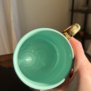 Puede incluir: Una taza de cer&aacute;mica turquesa con un asa dorada. El interior de la taza es de un tono turquesa m&aacute;s claro, con patrones de remolino visibles. El asa tiene un acabado dorado texturizado con detalles florales.