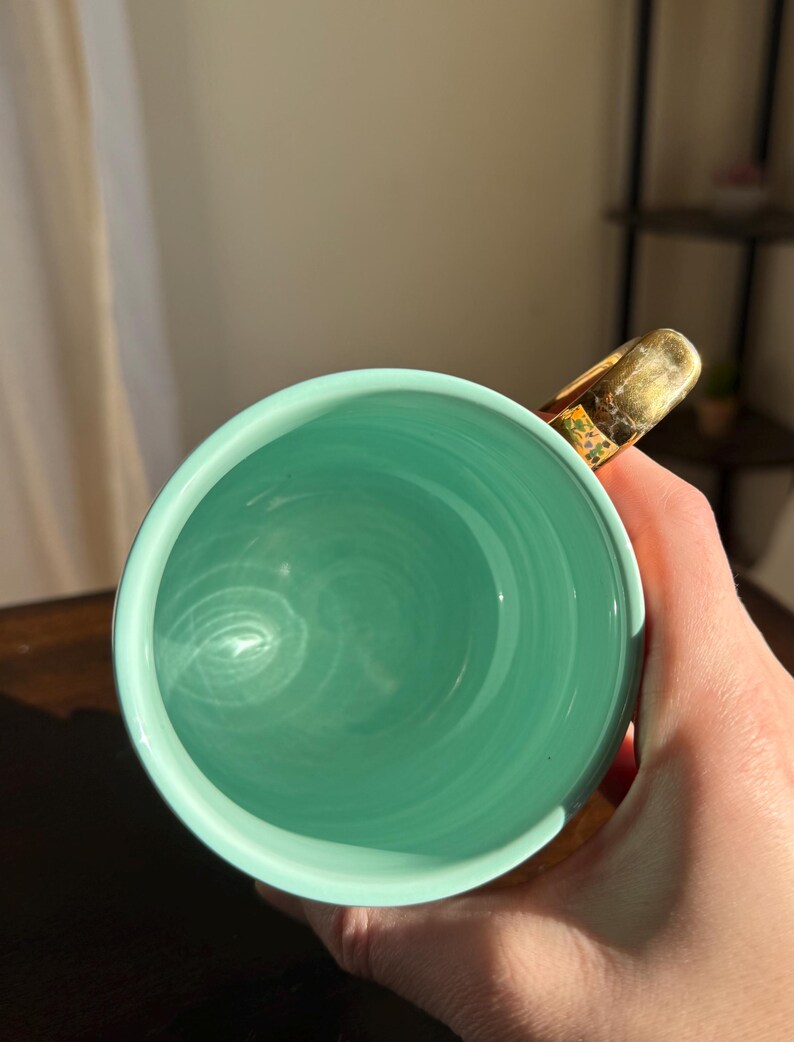 Puede incluir: Una taza de cer&aacute;mica turquesa con un asa dorada. El interior de la taza es de un tono turquesa m&aacute;s claro, con patrones de remolino visibles. El asa tiene un acabado dorado texturizado con detalles florales.