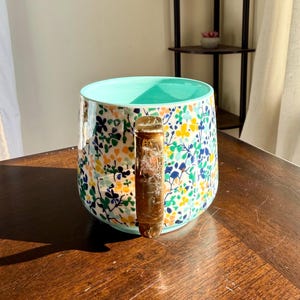 Puede incluir: Una taza de cer&aacute;mica con un estampado floral en amarillo, azul y verde sobre un fondo blanco. El interior es de color turquesa claro. El asa es de color &aacute;mbar transl&uacute;cido. La taza est&aacute; sobre una superficie de madera marr&oacute;n oscuro.