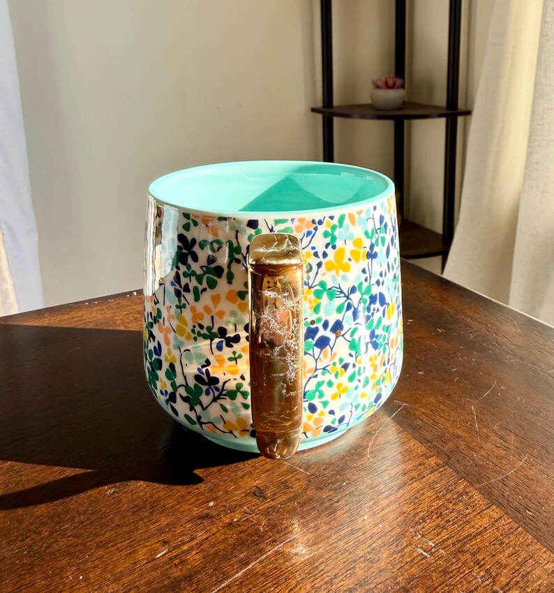 Puede incluir: Una taza de cer&aacute;mica con un estampado floral en amarillo, azul y verde sobre un fondo blanco. El interior es de color turquesa claro. El asa es de color &aacute;mbar transl&uacute;cido. La taza est&aacute; sobre una superficie de madera marr&oacute;n oscuro.