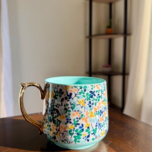 Puede incluir: Una taza de cer&aacute;mica con un estampado floral en tonos azules, verdes, amarillos y naranjas. La taza tiene un asa dorada y un interior azul claro. Est&aacute; sobre una superficie de madera.