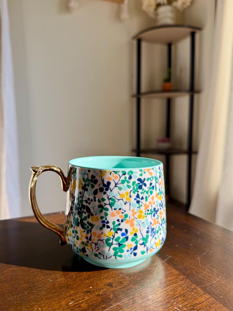 Puede incluir: Una taza de cer&aacute;mica con un estampado floral en tonos azules, verdes, amarillos y naranjas. La taza tiene un asa dorada y un interior azul claro. Est&aacute; sobre una superficie de madera.