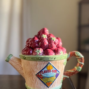 May include: A decorative ceramic teapot, designed as a watering can, filled with strawberries. The teapot features a wooden effect, a green trim, and a vine-like handle. The label reads "FLAVOR-SUM FANCY FRUIT".