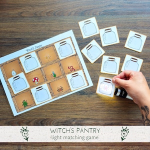 Op de afbeelding: Een Witch's Pantry licht-matching spel met een rechthoekig bord met rastervakken met illustraties van potten, een paddenstoel, een spin, een kever en een worm. Verschillende vierkante kaarten met potten zijn inbegrepen voor het spel.