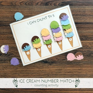 May include: A counting activity featuring a white card with the text "I CAN COUNT TO 5" and illustrated ice cream cones. Each cone has a different number of scoops. Colorful ice cream scoops are scattered around the card. The words "ICE CREAM NUMBER MATCH" are at the bottom.