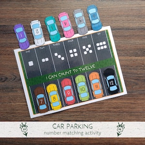 May include: A colorful car parking number matching activity. The image shows a game with numbered toy cars and a parking lot-themed game board. The board has the text "I CAN COUNT TO TWELVE" and the numbers one through twelve. The cars are various colors.