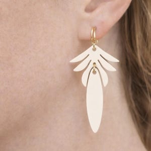 Può includere: Un orecchino color oro dal design unico. L'orecchino presenta un piccolo cerchio che si collega a un ciondolo a forma di foglia, color crema e stratificato. Il ciondolo ha una parte superiore a forma di palma e un fondo ovale e lungo. L'orecchino è indossato sull'orecchio di una persona.