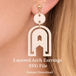 Op de afbeelding: Crèmekleurige gelaagde boogvormige oorbellen met goudkleurige accenten. De oorbellen hebben een geometrisch ontwerp met een boogvorm en een cirkelvormige bovenkant. De tekst "Layered Arch Earrings SVG File Instant Download" is zichtbaar.