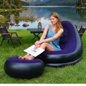 Puede incluir: Un sill&oacute;n inflable morado y negro con un reposapi&eacute;s a juego est&aacute; colocado sobre una zona de c&eacute;sped cerca de un lago. Una mujer est&aacute; sentada en la silla, sosteniendo un libro. Tambi&eacute;n se ve una peque&ntilde;a mesa negra y dos sillas plegables.