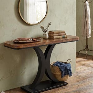 May include: A wooden console table with a dark brown top and black curved legs. A round gold mirror hangs above the table. A woven basket with a blue blanket sits below the table. Decorative items include books, a tray, and a vase.