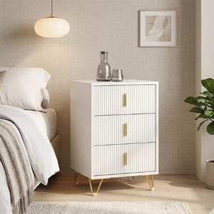 Könnte beinhalten: Ein weißer Nachttisch mit drei Schubladen, goldenen Griffen und Beinen. Die Schubladen haben ein vertikales Rillendesign. Eine Glasflasche und zwei Gläser stehen oben. Im Hintergrund ein Bett mit weißer Bettwäsche.