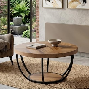 Op de afbeelding: Een ronde houten salontafel met een donker metalen onderstel. Het tafelblad heeft een visgraatpatroon en bevat een kleine kom en een boek. De tafel staat op een geweven vloerkleed. De achtergrond omvat een raam en een stoel.