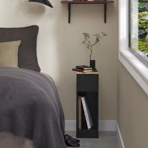 May include: A black bedside table with an open shelf and a closed top section. The top holds a small vase with a plant, notebooks, and a small wooden tray. Two books are in the open shelf. A bed with a gray blanket and pillows is in the background.