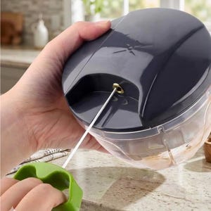 May include: A dark gray manual food chopper with a clear plastic base. A white cord extends from the top, connected to a green handle. The chopper is held in a hand, suggesting its use for food preparation.