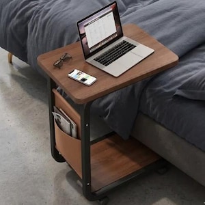 May include: A brown laptop table with a black frame and wheels. A laptop, smartphone, and glasses rest on the table's surface. A leather-like pocket holds magazines. The table is positioned next to a bed with gray bedding.