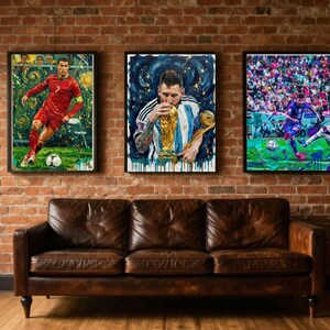 May include: Three framed rectangular wall art prints displayed above a brown leather sofa. The left print shows a soccer player in a red jersey, the center print shows a player kissing a gold trophy, and the right print shows a player on a field.