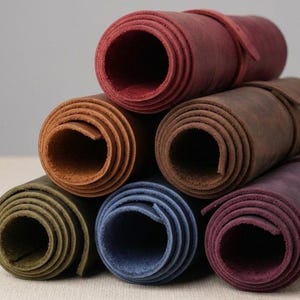 May include: A stack of rolled leather hides in various colors, including red, brown, blue, and green. Each roll is secured with a matching leather strap. The hides appear to be of high quality and suitable for crafting.