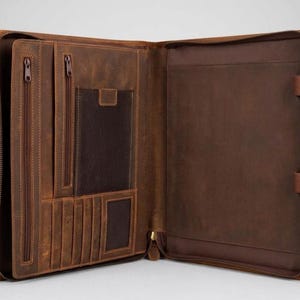 May include: Open brown leather portfolio with multiple pockets, card slots, and two zipped compartments. The portfolio is open, revealing a spacious interior and a smaller dark brown pocket. A gold-coloured zip runs around the perimeter.